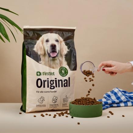 Förpackning med FirstVet Original hundfoder, en person häller foderkulor i en skål bredvid påsen. Bakgrunden inkluderar en grön växt och en blåvitrutig duk.