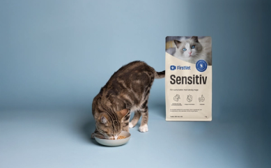 En brunfläckig katt som äter mat ur en matskål. Snett bakom står en påse kattmat av FirstVet Sensitiv.