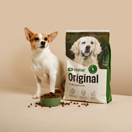 En förpackning med hundfoder från FirstVet märkt 'Original', anpassad för alla vuxna hundar. Förpackningen visar en glad golden retriever, och bredvid står en mindre hund intill en skål med torrfoder som har spillt ut några bitar. Texten på förpackningen nämner produktens egenskaper: hälsosam proteinkälla, omega-3 från fiskolja och prebiotika för en sund tarmflora.